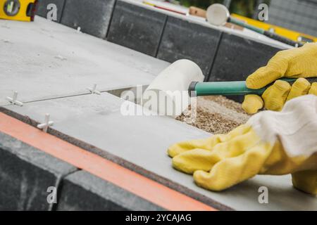 Un operaio edile che indossa guanti gialli livella le finitrici di pietra utilizzando un mazzuolo in gomma, garantendo precisione in un progetto di architettura paesaggistica all'aperto. Foto Stock