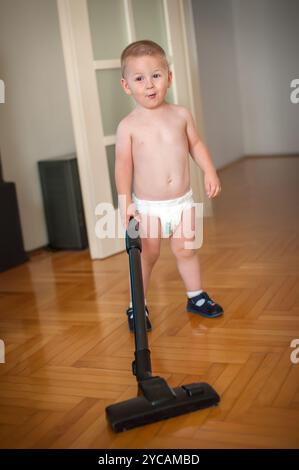 Un bambino in un pannolino si trova in un soggiorno illuminato e tiene in mano un aspirapolvere. Il bambino ha un aspetto gioioso mentre è impegnato ad aiutare la casa Foto Stock