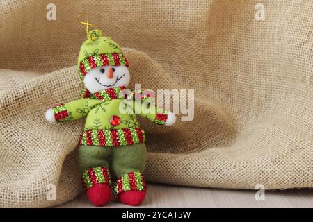 Pupazzo di neve in abito verde davanti allo sfondo in tessuto di iuta per le festività natalizie invernali di dicembre. Allegro carattere di peluche, copia spazio. Foto Stock