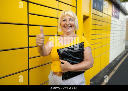 Donna anziana degli anni '50 che mostra il pollice su come ottenere il pacco dalla cella del terminale postale automatico all'aperto. Concetto di delivery senza contatto e intelligente. Idea di Foto Stock