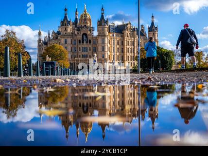 22 ottobre 2024, Meclemburgo-Pomerania occidentale, Schwerin: Passanti a piedi lungo un sentiero di fronte al castello di Schwerin e si vedono riflessi in una pozzanghera. Con il sole e le temperature miti, il clima autunnale nella Germania settentrionale mostra il suo lato amichevole. Foto: Jens Büttner/dpa Foto Stock