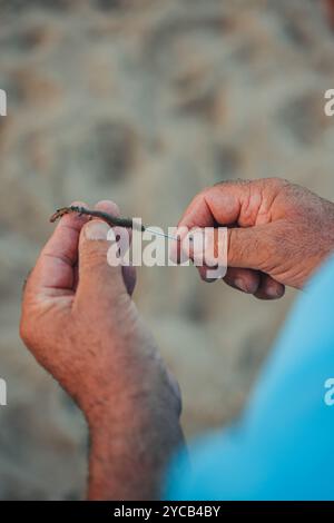 Immagine ravvicinata delle mani di un pescatore anziano che infilano meticolosamente un verme su un gancio, catturato su uno sfondo sabbioso sfocato a Valencia, in Spagna Foto Stock