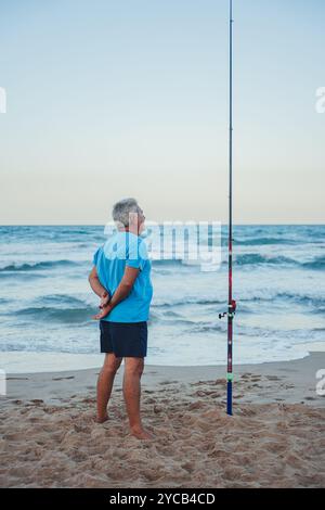 Un uomo anziano si trova sulle coste sabbiose di Valencia, in Spagna, osservando le onde dell'oceano mentre pesca al tramonto la canna da pesca è posta in rilievo ne Foto Stock