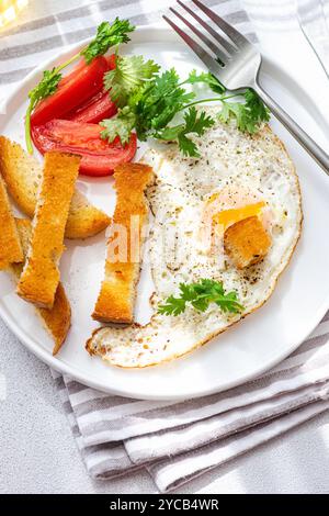 Piatto bianco con un pasto salutare con un uovo soleggiato, fette di pomodoro fresche, bastoncini di pane croccanti e una spolverata di prezzemolo fresco. Ideale per un Foto Stock