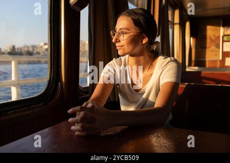 Una donna premurosa guarda lontano dalla macchina fotografica attraverso una finestra del traghetto, crogiolandosi nella calda luce dorata mentre attraversa il Bosforo di Istanbul. La S Foto Stock