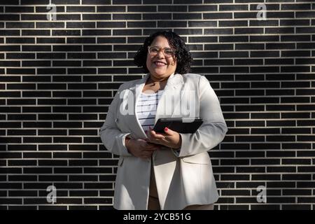 Donna ispanica professionale con occhiali e blazer bianco, sorride alla fotocamera mentre tiene in mano un tablet davanti a una parete di mattoni scuri. Foto Stock