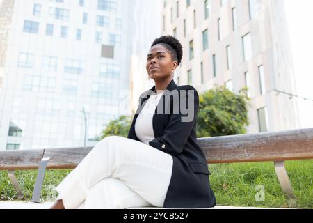 Una donna d'affari afroamericana guarda con fiducia mentre siede su una panchina di legno del parco, circondata da grattacieli e vegetazione urbana. Foto Stock