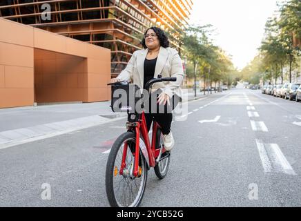 Una donna ispanica adulta si diverte a fare un piacevole giro in bicicletta su una tranquilla strada cittadina, guardando in lontananza con un'espressione contemplativa. Foto Stock