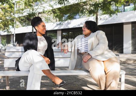 Due donne professionali, una ispanica e l'altra afroamericana, si impegnano in una conversazione vivace mentre sono sedute su una panchina del parco in un ambiente urbano. Foto Stock