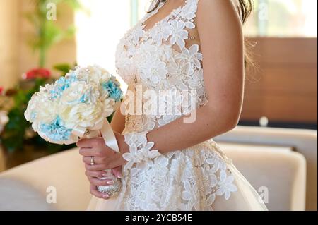 Primo piano di una sposa in un dettagliato abito da sposa in pizzo, che regge con grazia un bouquet di fiori blu e bianco Foto Stock