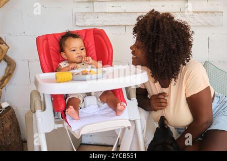 Una madre afroamericana sorride amorevolmente al suo bambino seduto su un seggiolone rosso. Il bambino, anche afroamericano, sembra curiosamente mentre tiene in mano una spiga Foto Stock