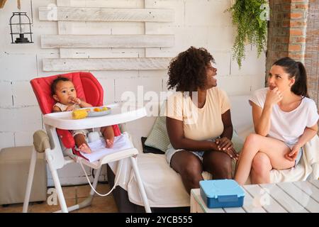 Una gioiosa scena familiare multietnica con una donna afroamericana, una donna caucasica e il loro bambino di razza mista che si gusta un pasto. Il bambino è seduto in un Foto Stock