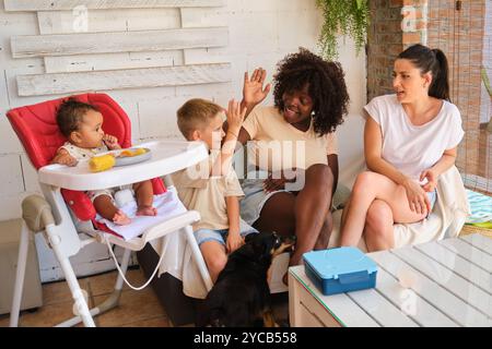 Una famiglia multirazziale è riunita intorno a un tavolino. La donna afroamericana dà un cinque alto a un ragazzo caucasico, mentre una donna ispanica parla Foto Stock