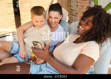 Una famiglia allegra e multirazziale trascorre momenti di qualità all'aperto. Una donna afro-americana e una donna caucasica, entrambe guardano uno smartphone, ridono e si divertono Foto Stock