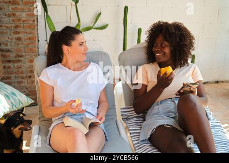 Una donna caucasica e una donna afroamericana, entrambe vestite casualmente, sono sedute su un patio. La donna caucasica guarda la donna afroamericana Foto Stock