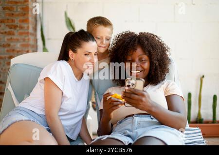 Una gioiosa famiglia multietnica si riunisce in un accogliente soggiorno. Una donna afroamericana, una donna caucasica e un ragazzo caucasico si impegnano felicemente con uno smartph Foto Stock