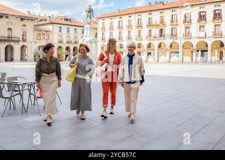Quattro donne mature in abiti casual camminano allegramente attraverso una spaziosa piazza della città, con edifici storici e una statua sullo sfondo Foto Stock
