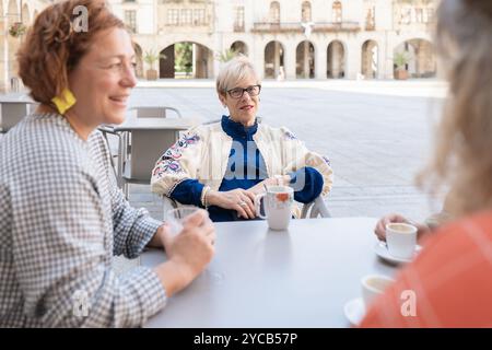 Tre donne anziane, sorridenti e impegnate nella conversazione, gustate una tazza calda di caffè insieme in un caffè all'aperto in una piazza della città Foto Stock