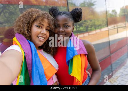 Due giovani donne, una afroamericana e un'altra latinoamericana, sorridono gioiosamente in un parco vestito con abbigliamento casual e drappeggiato con bandiera arcobaleno Foto Stock
