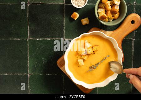 Una sontuosa porzione di minestra cremosa di zucca guarnita con timo, crostini al pepe, Un cucchiaio di legno e porzioni extra di condimenti aggiungono un'atmosfera accogliente e casalinga Foto Stock