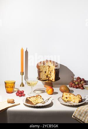 Un tavolo arredato con gusto che mette in mostra il tradizionale panettone italiano, circondato da frutta fresca, uva, una mela ed eleganti bicchieri, immersi in so Foto Stock