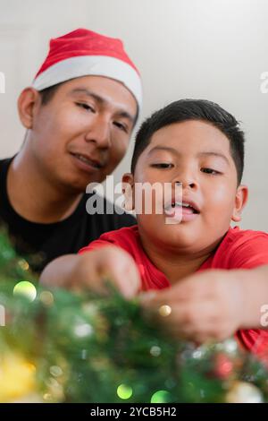 Un'emozionante scena di un padre latino e del suo giovane figlio che indossano un cappello di Babbo Natale mentre decorano un albero di Natale, condividendo un gioioso momento familiare Foto Stock