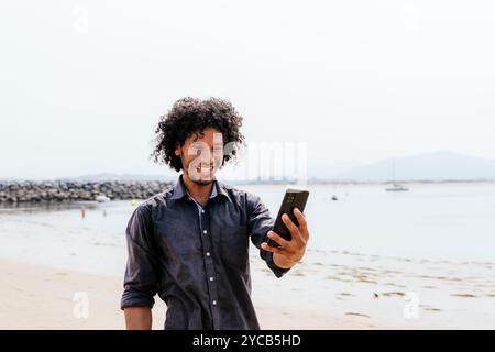 Un uomo allegro con i capelli ricci ama una giornata in spiaggia, catturando un momento con un selfie con il suo smartphone, mostrando un'uscita casual e gioiosa Foto Stock