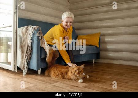Una donna matura allegra con i capelli biondi corti, vestita con un maglione giallo, accarezza un gatto di zenzero grande in un ambiente accogliente caratterizzato da un arredamento moderno e. Foto Stock