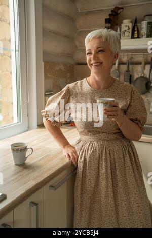 Una donna allegra e matura con i capelli grigi corti, in piedi in una cucina rustica, sorridente mentre regge una tazza di caffè. La luce entra da una finestra, gettando Foto Stock