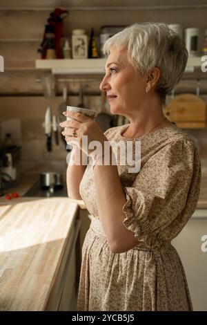 Vista laterale di una donna matura e serena, con i capelli grigi corti, che sorseggia un caffè e guarda attentamente in un ambiente rustico da cucina. Indossa una dres floreale Foto Stock