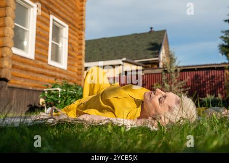 Una donna matura con un vestito giallo vivace gode di un momento di pace, sdraiata sull'erba di fronte a una casa rustica. I suoi occhi sono chiusi mentre abbraccia Foto Stock