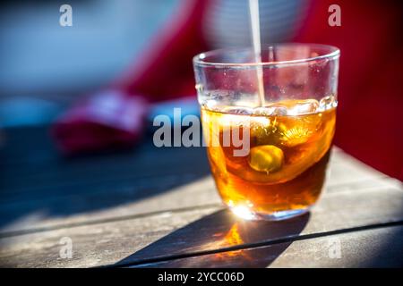 Un primo piano di un rinfrescante bicchiere di vermouth con ghiaccio e guarnizione di olive. La luce del sole esalta i colori vivaci, creando un'atmosfera invitante Foto Stock