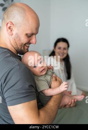 In primo piano sorride papà con neonato in braccio. Mamma si siede a letto e guarda le persone care. Al centro dell'attenzione c'è l'uomo. Famiglia felice. Foto Stock