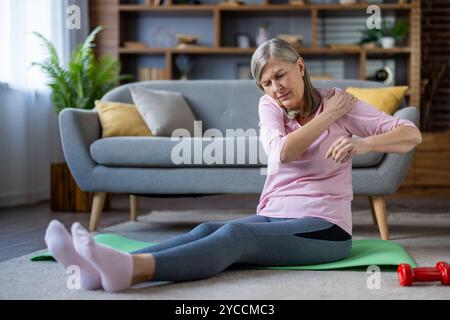 Donna anziana con dolore alle spalle siede sul tappetino da yoga in salotto. Signora anziana in camicia rosa e pantaloni da yoga si sente disagio dopo l'allenamento, illustratin Foto Stock