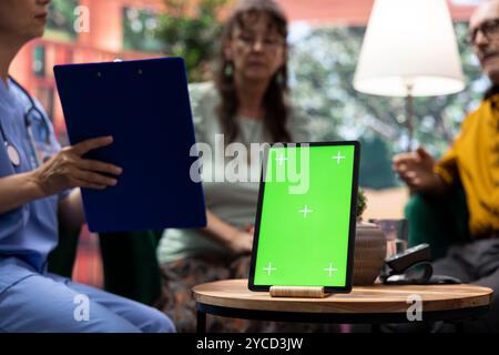 Uomo e donna anziani che parlano con l'infermiera a casa loro con uno schermo verde sul tablet, si divertono a chiacchierare insieme e chiedono indicazioni. La coppia di anziani si sente bene e si rilassa durante la visita di check-up. Foto Stock