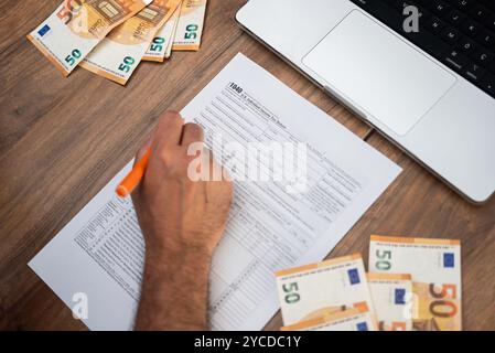 Il dirigente aziendale siede al tavolo con banconote in euro e laptop con penna in mano compilando il modulo 1040 Foto Stock