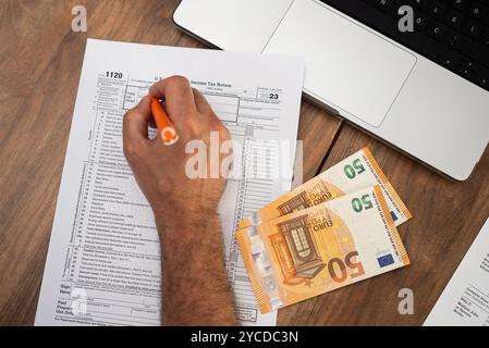 Il consulente fiscale compila meticolosamente il modulo fiscale 1120 sul tavolo utilizzando un computer portatile e facendo riferimento alle banconote in euro Foto Stock