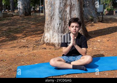 Bambino brasiliano seduto su un tappetino da yoga e che pratica respirazione nella classe all'aperto 1. Foto Stock