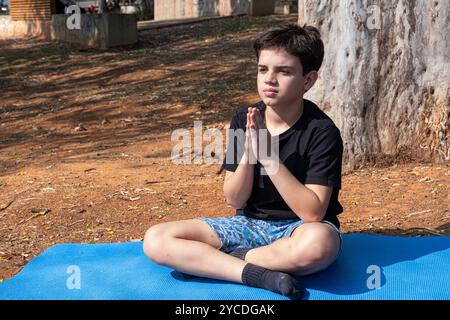 Bambino brasiliano seduto su un tappetino da yoga e che pratica respirazione nella classe all'aperto 2. Foto Stock