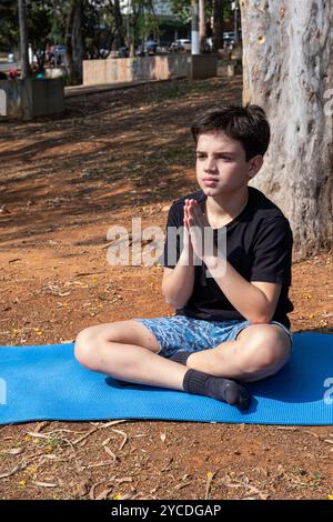 Bambino brasiliano seduto su un tappetino da yoga e che pratica respirazione nella classe all'aperto 3. Foto Stock