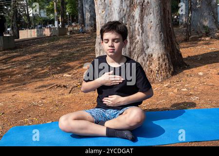 Bambino brasiliano seduto su un tappetino da yoga con le mani sul petto e sull'addome nella classe all'aperto 4. Foto Stock