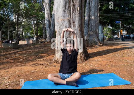 Bambino brasiliano seduto su un tappetino da yoga e che si allunga nella classe all'aperto 6. Foto Stock