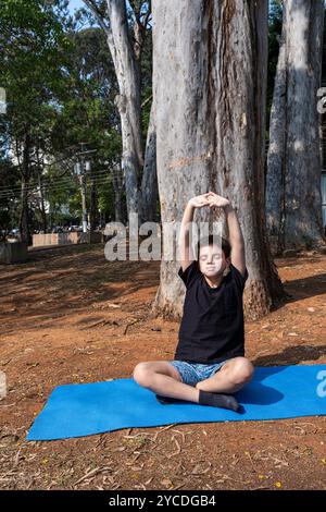 Bambino brasiliano seduto su un tappetino da yoga e che si allunga nella classe all'aperto 7. Foto Stock