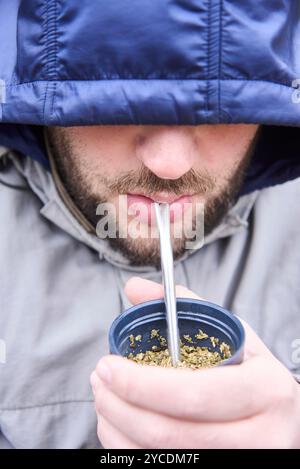 Ritratto di un giovane uomo argentino che beve un compagno, una bevanda calda tradizionale, in una fredda mattina d'inverno. Composizione da vicino, cappuccio della giacca Foto Stock