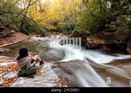 Escursionista seduto vicino alla cascata che scorre sul fiume Looking Glass Creek in autunno - Pisgah National Forest, vicino a Brevard, North Carolina, Stati Uniti Foto Stock