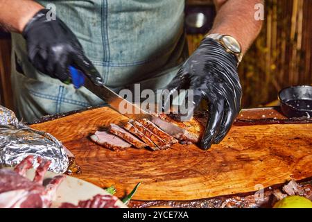 Uno chef con guanti neri affetta la carne appena cucinata su una tavola di legno, preparando un piatto saporito con precisione. Foto Stock