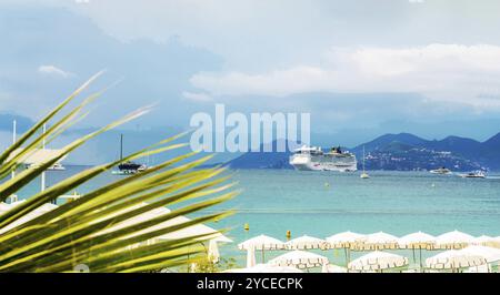 Splendida vista sulla baia del resort di Cannes (Mar Mediterraneo) con una grande nave da crociera. e piccole barche a vela. Foto di Taling dalla Croisette: Ombrelloni, esterno Foto Stock