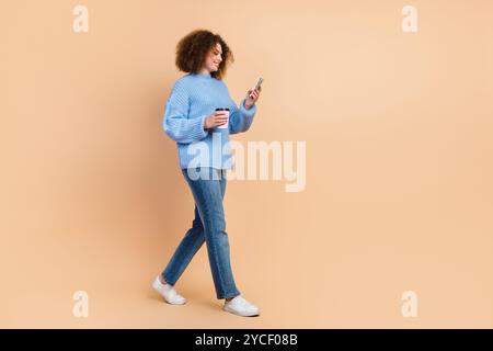 Foto di una giovane ragazza latina in stile casual con tazza di caffè in carta ecologica. Ordina ciambelle extra utilizzando lo smartphone isolato su sfondo di colore beige Foto Stock