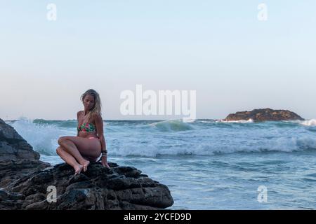 Donna bionda con capelli sciolti vestita in bikini seduta su una scogliera di fronte al mare bagnata dall'acqua delle onde Foto Stock