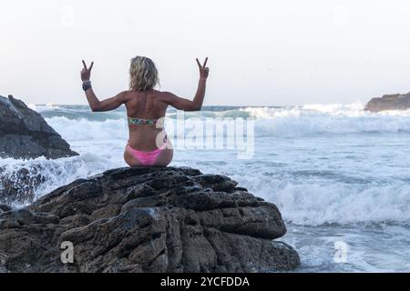 Donna bionda con capelli sciolti vestita in bikini seduta su una scogliera di fronte al mare bagnata dall'acqua delle onde Foto Stock